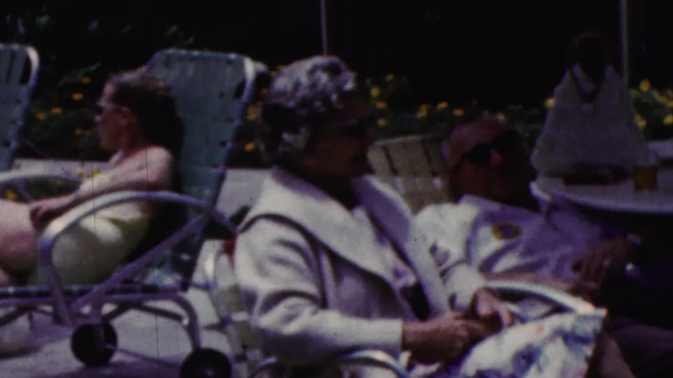 California 1961 home-movie frame showing women in light jackets and sunglasses seated on patio furniture near lounge chairs, with garden planting in the background.