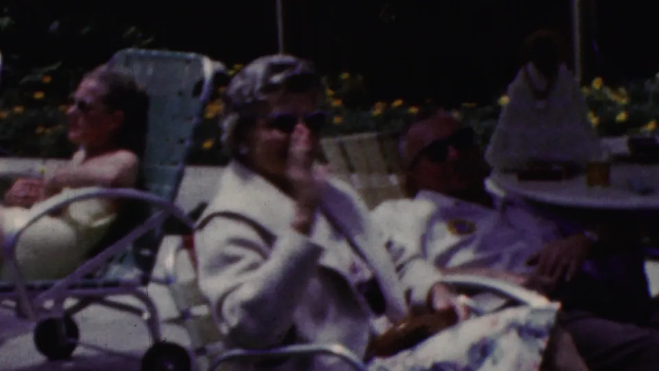 California 1961 home-movie frame showing older adults seated outdoors beside a patio table and lounge chairs, including a woman in sunglasses facing the camera.