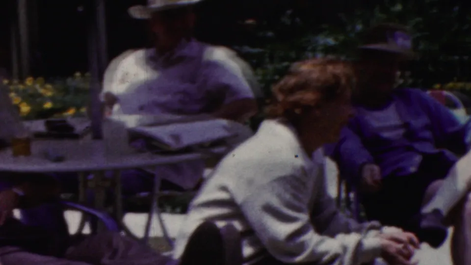 California 1961 home-movie frame showing adults gathered outdoors around a patio table under an umbrella, with a hat-wearing man and a seated woman in light clothing visible.