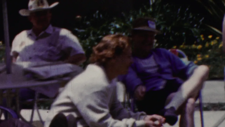 California 1961 home-movie frame showing adults lounging outdoors near patio furniture, including a woman in a light sweater and seated men in hats or casual shirts.