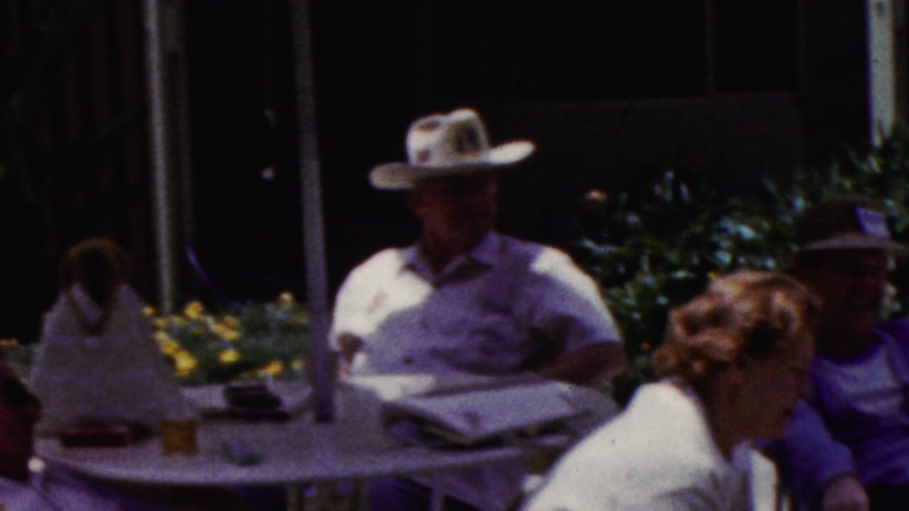 California 1961 home-movie frame with adults seated at a shaded patio table, including a man in a broad-brimmed hat near a newspaper and a woman turned toward him.