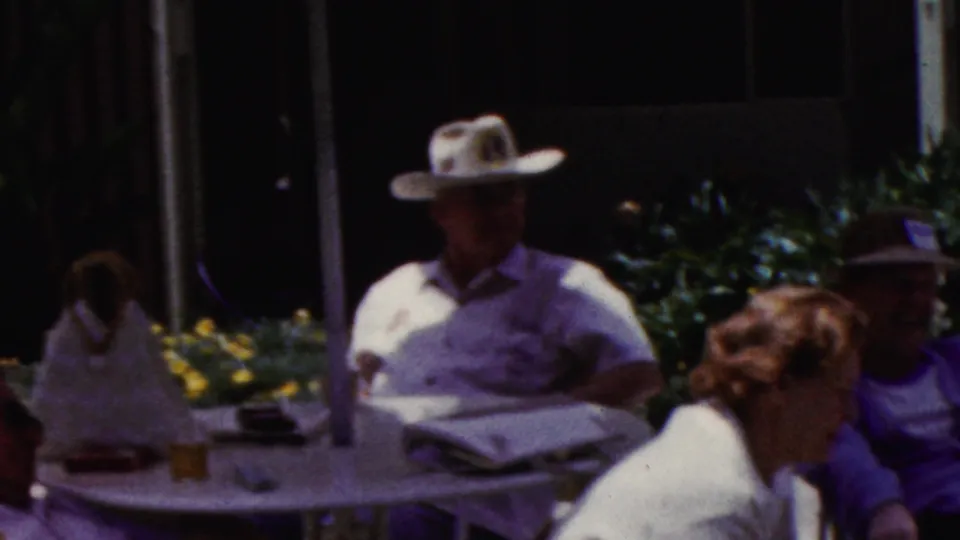 California 1961 home-movie frame with adults seated at a shaded patio table, including a man in a broad-brimmed hat near a newspaper and a woman turned toward him.