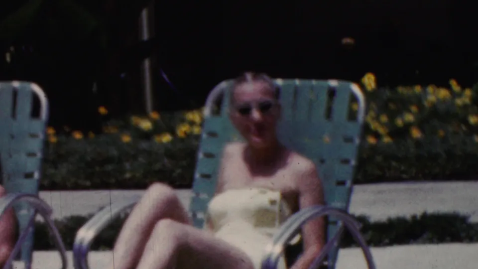 California 1961 home-movie frame showing an adult woman in sunglasses and a yellow swimsuit reclining on a turquoise lounge chair near flowers and a paved outdoor area.