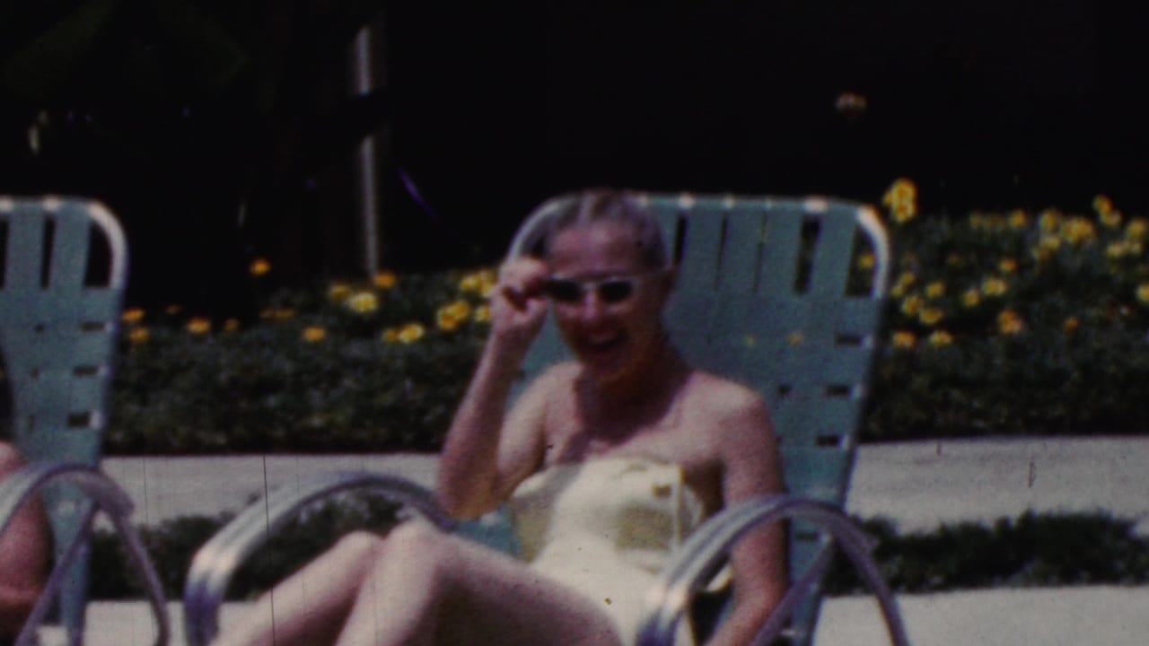 1961 California home-movie frame showing women relaxing by a poolside area on turquoise lounge chairs, including a smiling woman in sunglasses and a light swimsuit.
