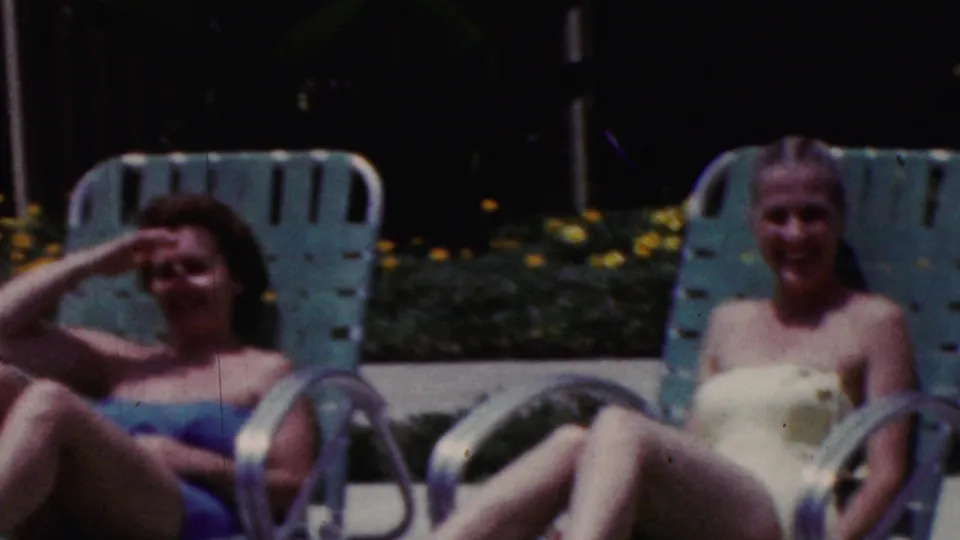 1961 California poolside frame with a woman in a blue swimsuit reclining on a turquoise lounge chair while raising her hand against the sunlight.
