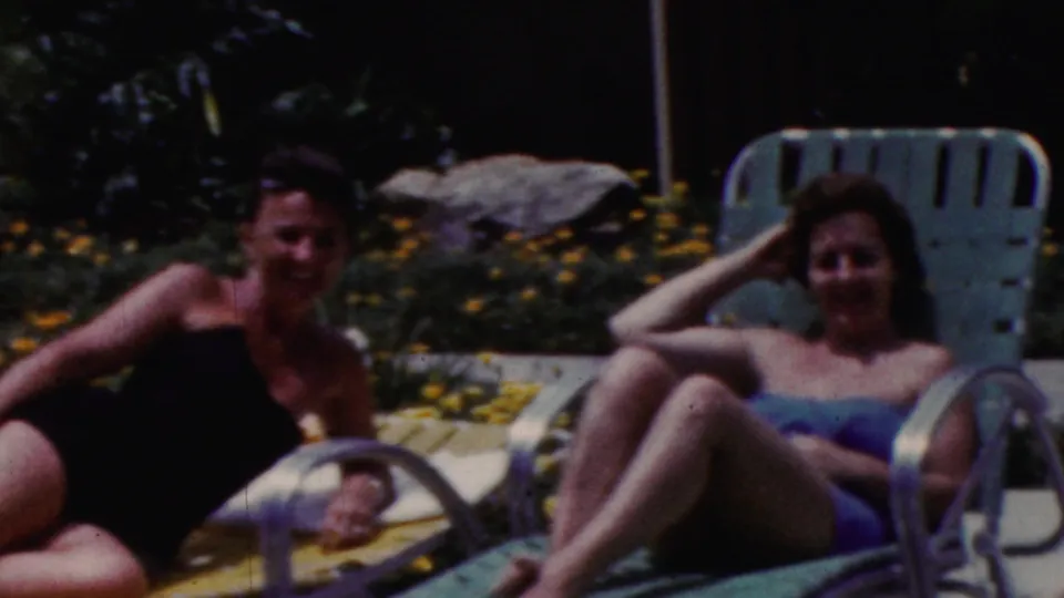1961 California home-movie frame showing two women by turquoise poolside lounge chairs, one in a dark swimsuit and one in a blue swimsuit smiling toward the camera.