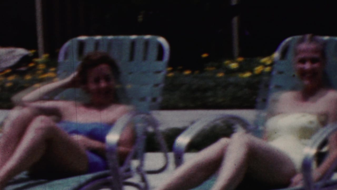 Two women in swimsuits reclining on green poolside loungers in bright sun.