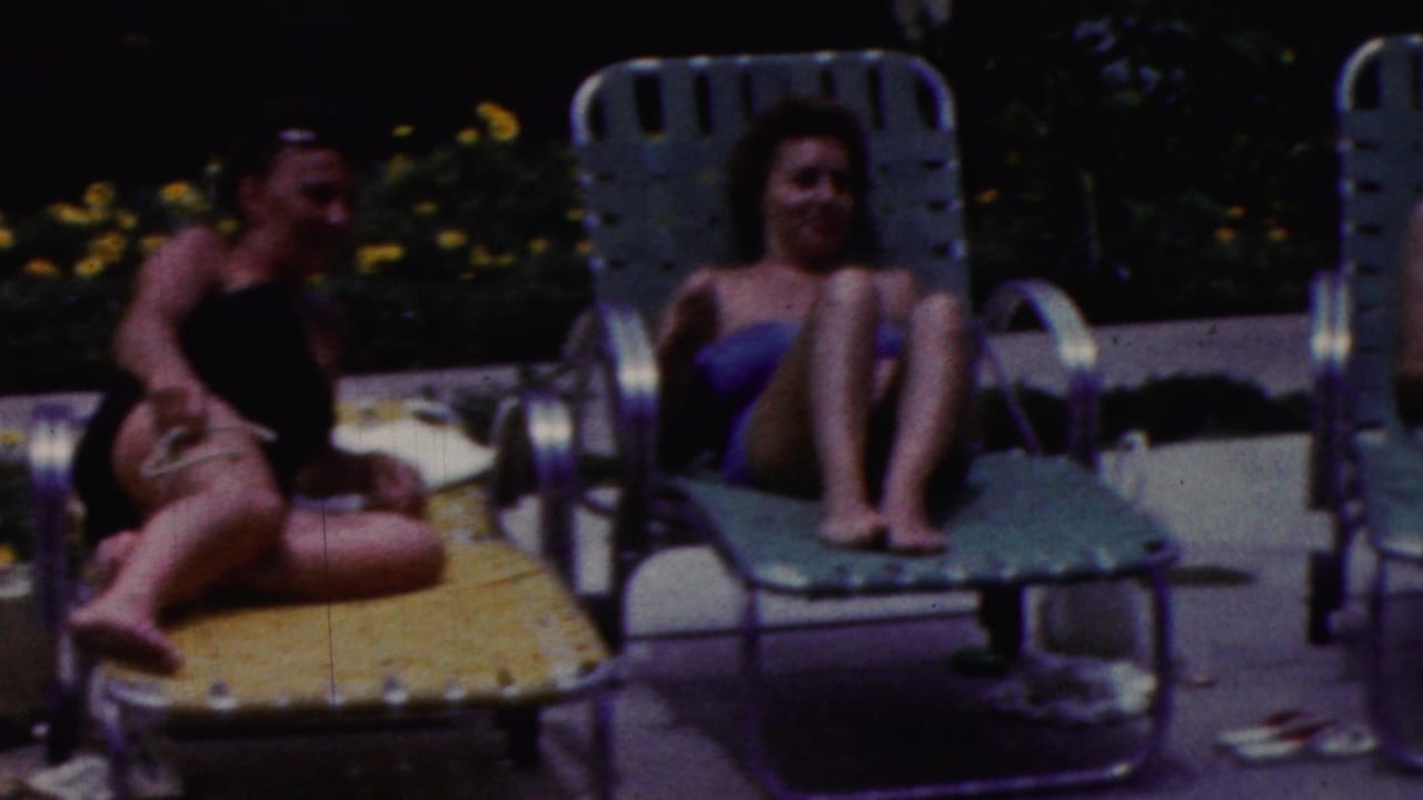 Two women sit on green poolside loungers in 1961 California home-movie footage, with a third adult partly visible on a yellow chaise between them.