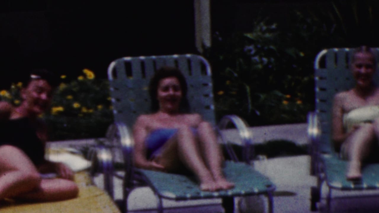 In supported 1961 California home-movie footage, two women in bathing suits sit on green lounge chairs by a poolside patio with garden planting behind them.