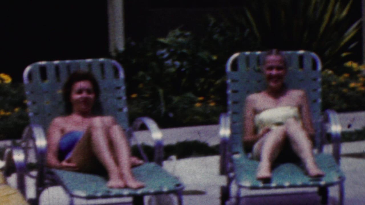 Two women recline and smile on green metal lounge chairs in 1961 California poolside home-movie footage, with dark foliage and yellow flowers in the background.