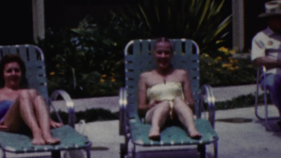 A woman in a light bathing suit sits on a green lounge chair, with another seated woman partly visible at left and a standing adult partly visible behind the chairs.
