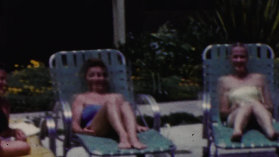 Two women sit facing the camera on green lounge chairs in supported 1961 California home-movie footage, framed by garden foliage and a poolside patio.