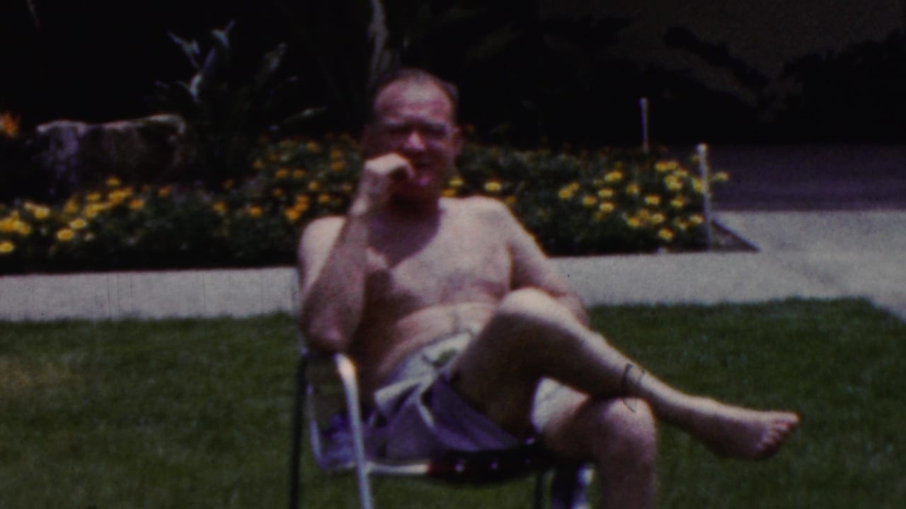 In 1961 California home-movie footage, a shirtless adult man in swim trunks sits barefoot in a folding lawn chair beside a lawn and flower bed.