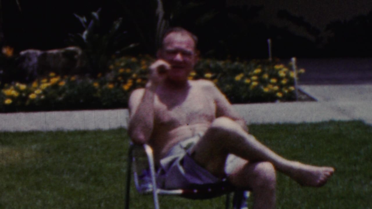 Man in swim trunks seated on a lawn chair with flowers and plants behind him.