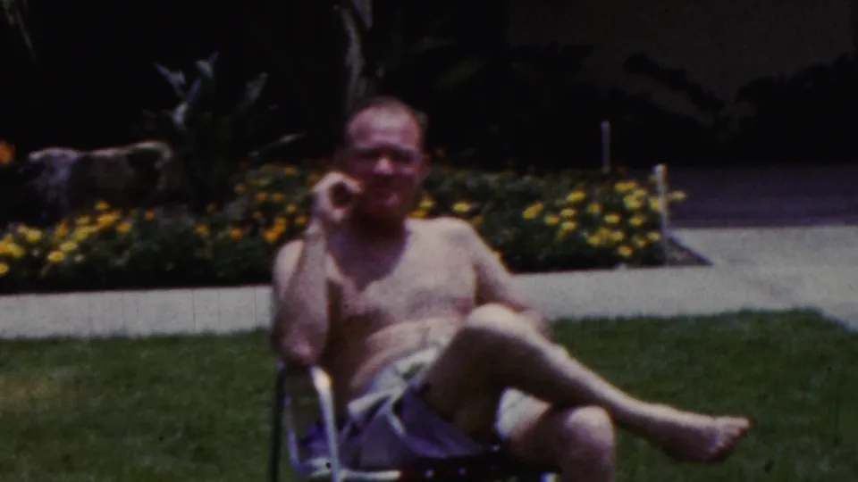A soft 8mm frame shows the man leaning in the chair on the grass, with the concrete walkway and planted border visible behind him.