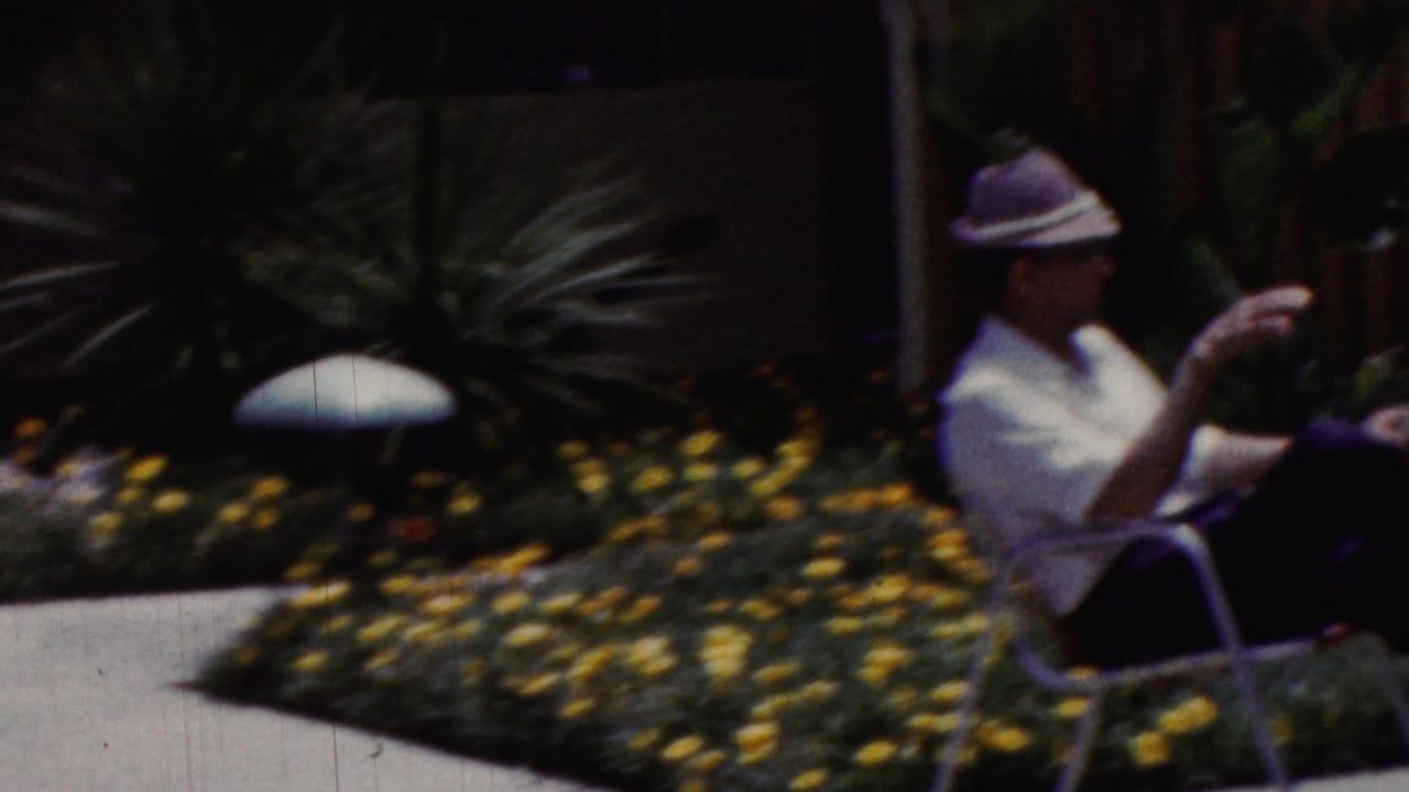 In 1961 California home-movie footage, an adult wearing a light-colored hat and white shirt sits on a patio chair near yellow flowers and desert-style plants.