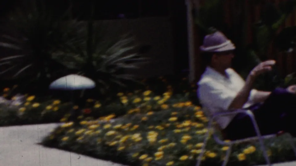 In 1961 California home-movie footage, an adult wearing a light-colored hat and white shirt sits on a patio chair near yellow flowers and desert-style plants.