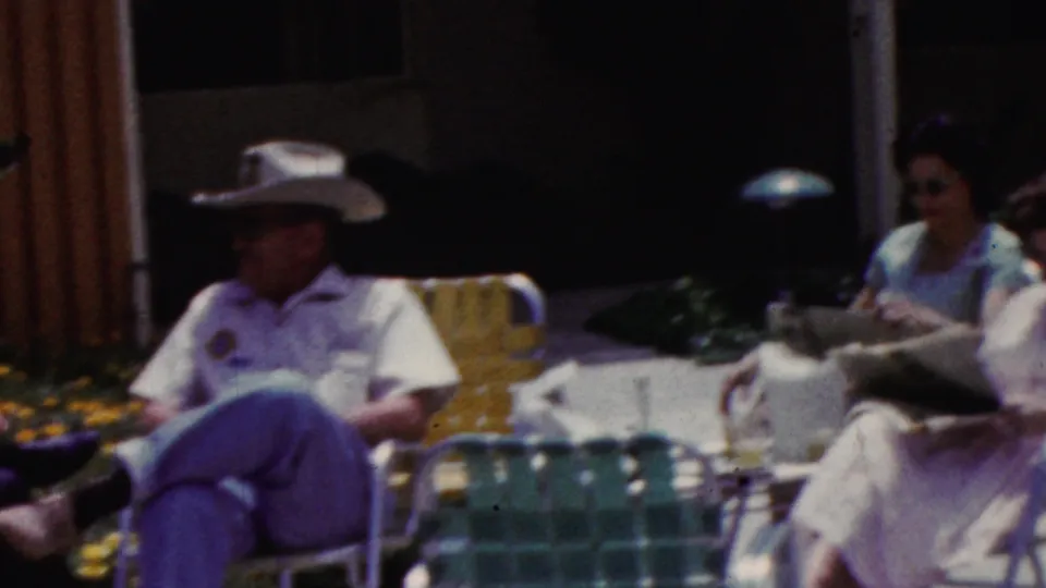 Adults sit together on a sunny patio in 1961 California footage, including a person in a light hat and another figure holding a newspaper near green lawn chairs.