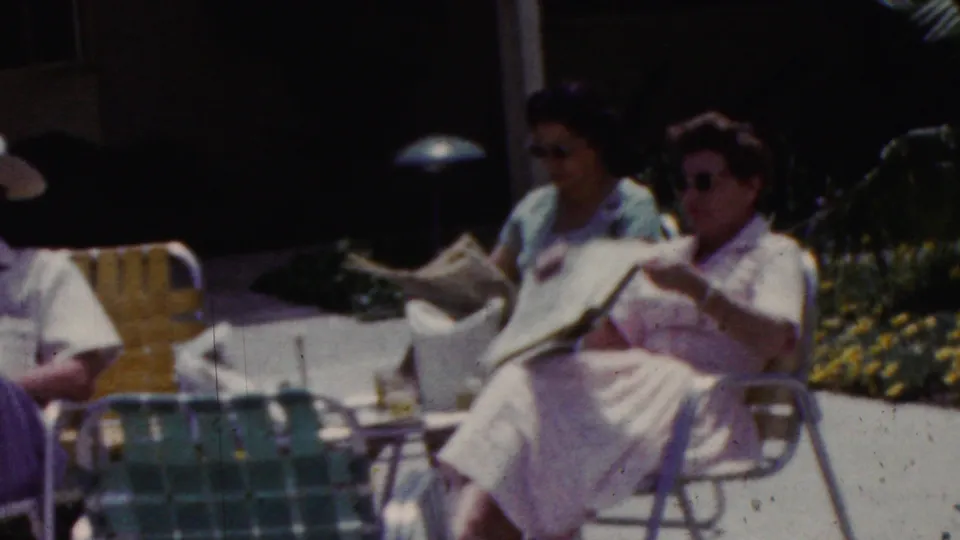 Two women in dresses sit in metal lawn chairs on a sunny California patio, with one reading a newspaper and garden flowers visible nearby.