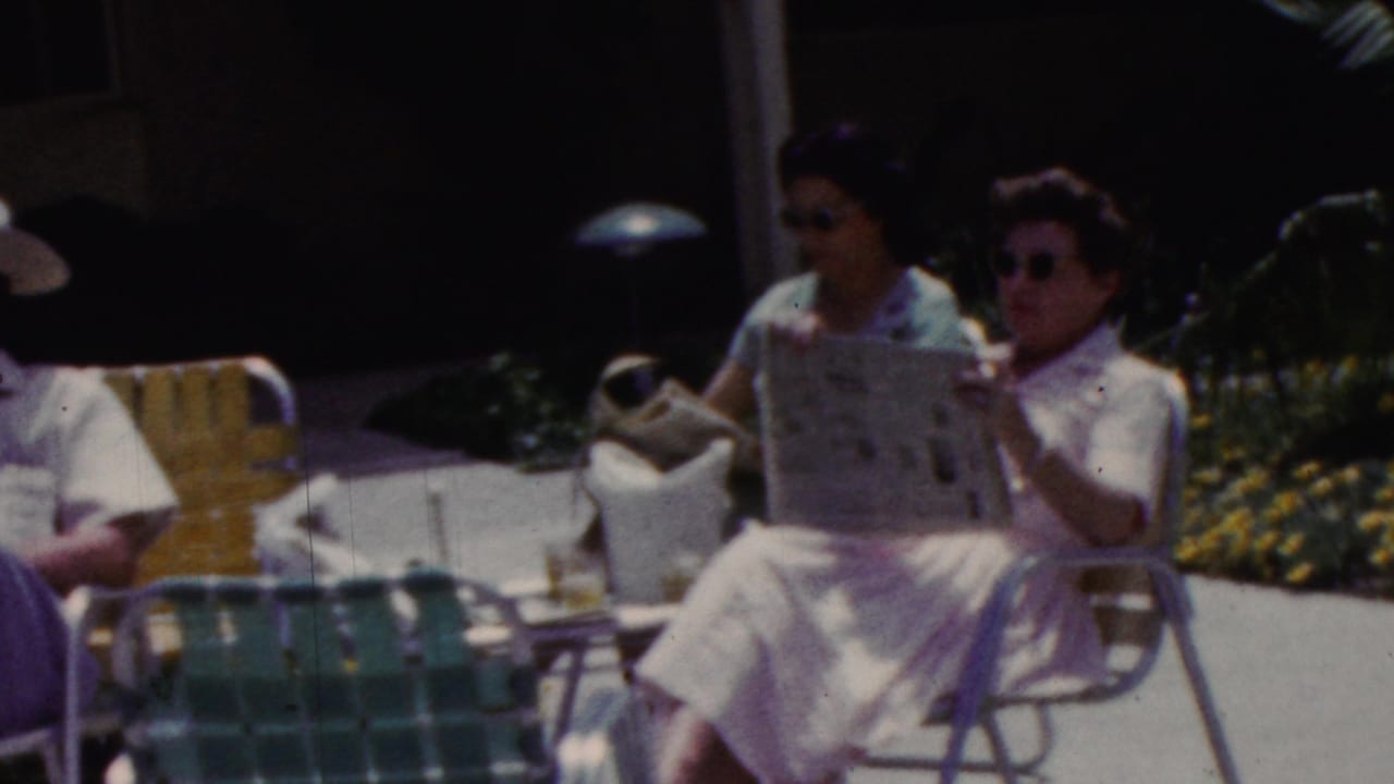 Two women in white clothing and sunglasses sit outdoors in California, 1961 metadata, reading newspapers beside metal lawn chairs and patio furniture.