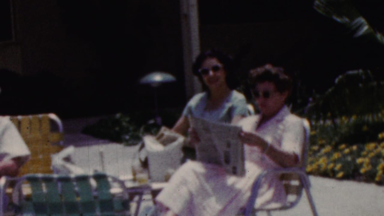 A home-movie frame shows two women seated on a patio in lawn chairs, with one holding an open newspaper and flowers and shaded building areas behind them.