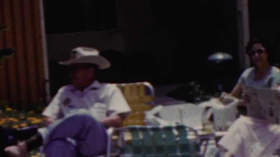 A man wearing a cowboy hat sits in a lawn chair near a woman with a newspaper, with patio seating, flowers, and a building facade visible in the background.