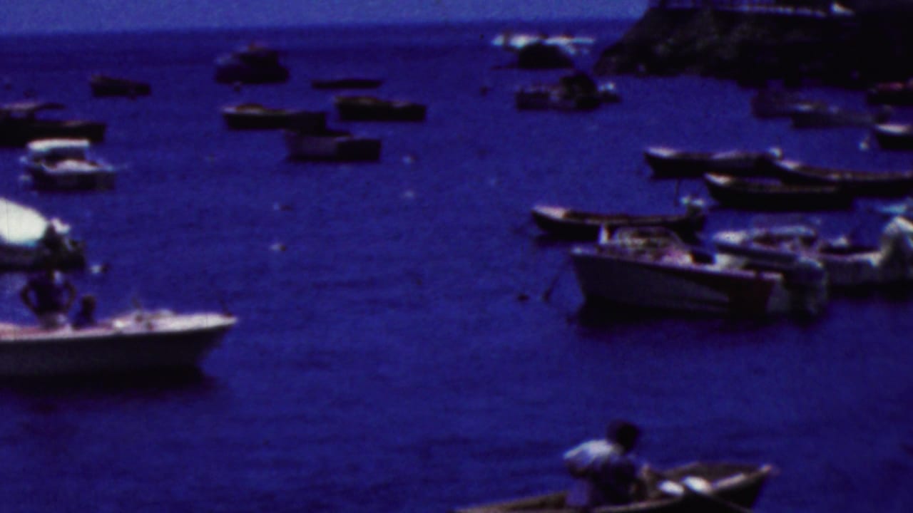 1961 California home-movie frame showing a harbor with small boats anchored across deep blue water and a person rowing close to the camera position.