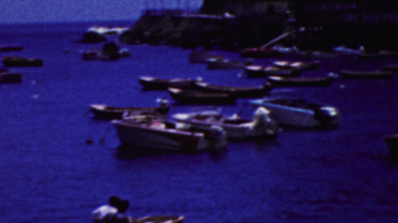 Anchored boats float near a rocky shoreline and waterside buildings in blue-tinted harbor water.
