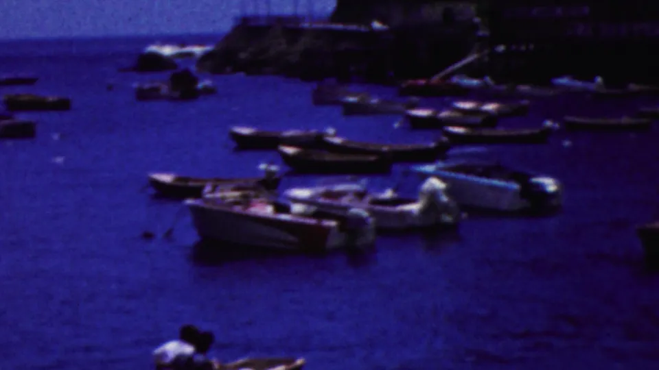 1961 California coastal harbor view with several small boats moored in blue water, rocky shore beyond, and simple waterfront structures in the background.