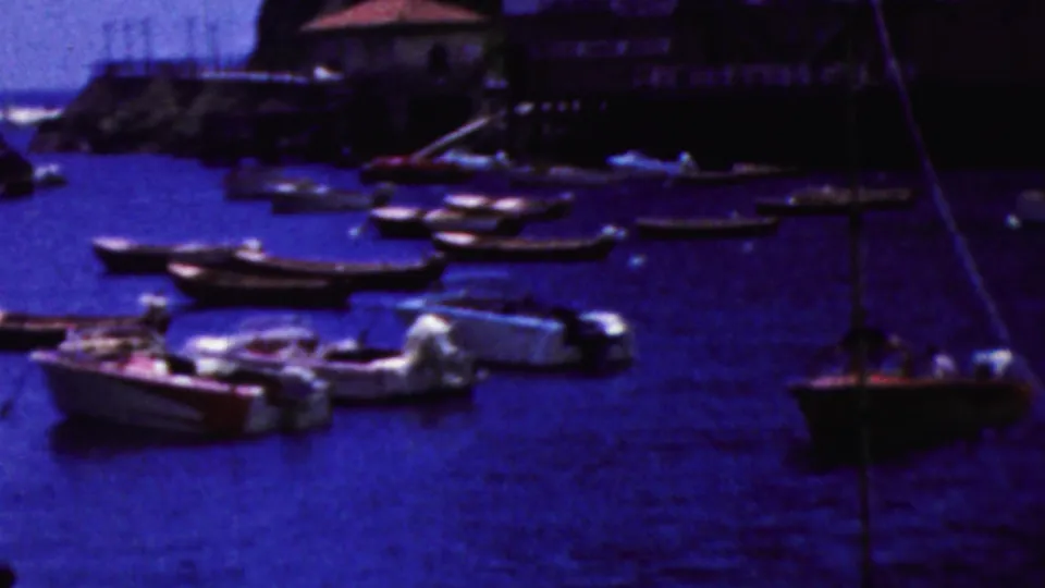 1961 California harbor frame showing clustered small boats, a prominent mooring line in the foreground, and simple shoreline buildings above the water.