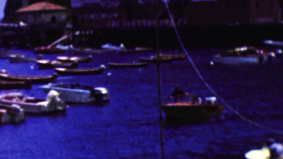 1961 California home-movie view of a small foreground boat with people aboard, moored boats around it, and a waterfront building along the rocky shore.