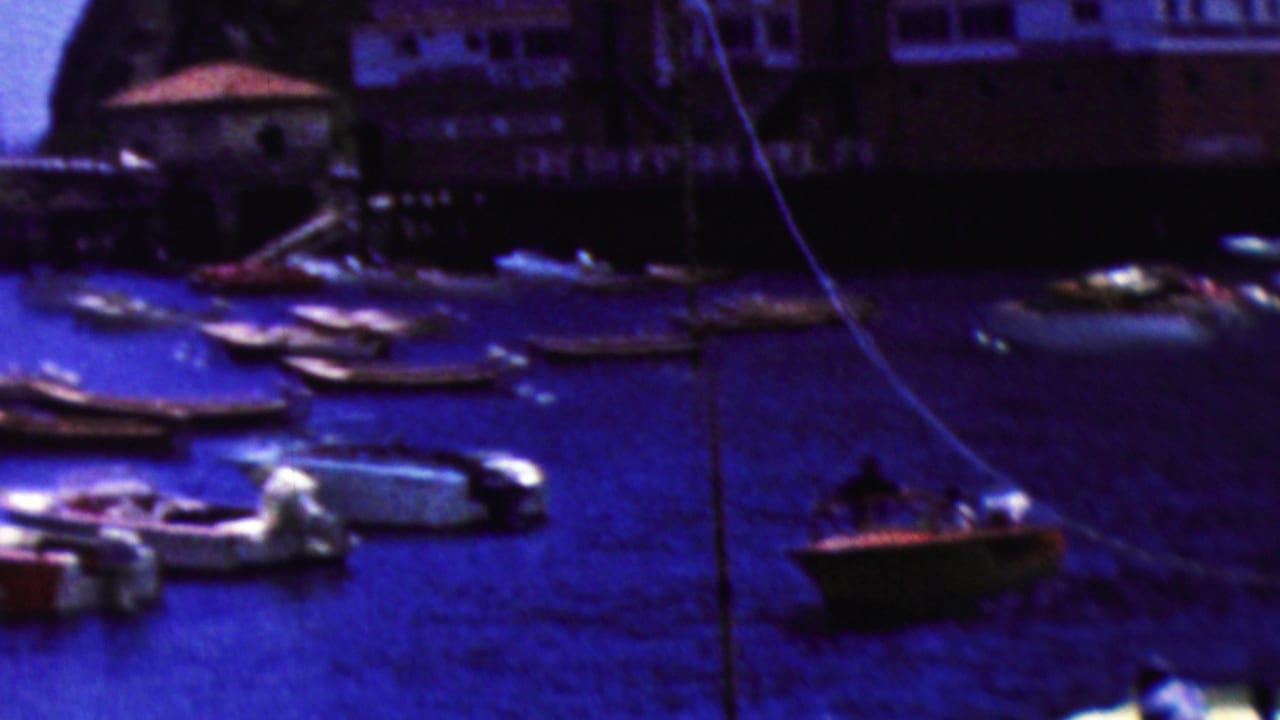 1961 California 8mm home-movie frame showing small boats scattered across deep blue harbor water near waterfront buildings and a rocky shoreline.