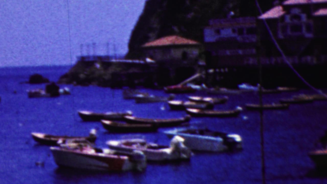 1961 California harbor view with moored small boats, a rocky outcrop, waterfront buildings, and hazy ocean horizon in soft 8mm color.