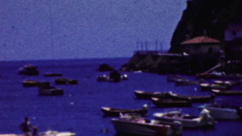 1961 California 8mm frame showing a busy small-boat harbor below a rocky cliff and waterfront structures, with a distant person aboard one boat.