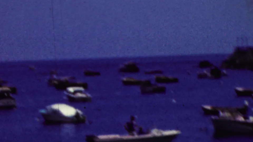 Soft 1961 California 8mm harbor frame with small boats spread across blue water near a distant shoreline and faint figures at the lower edge.