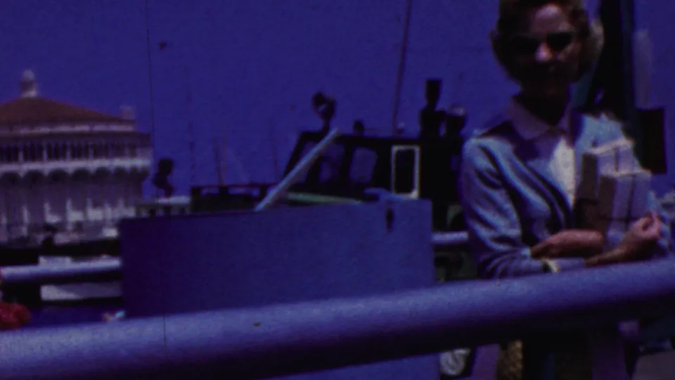The woman remains near the boat railing, holding items as a pale waterfront building and dark boat equipment appear in the background.