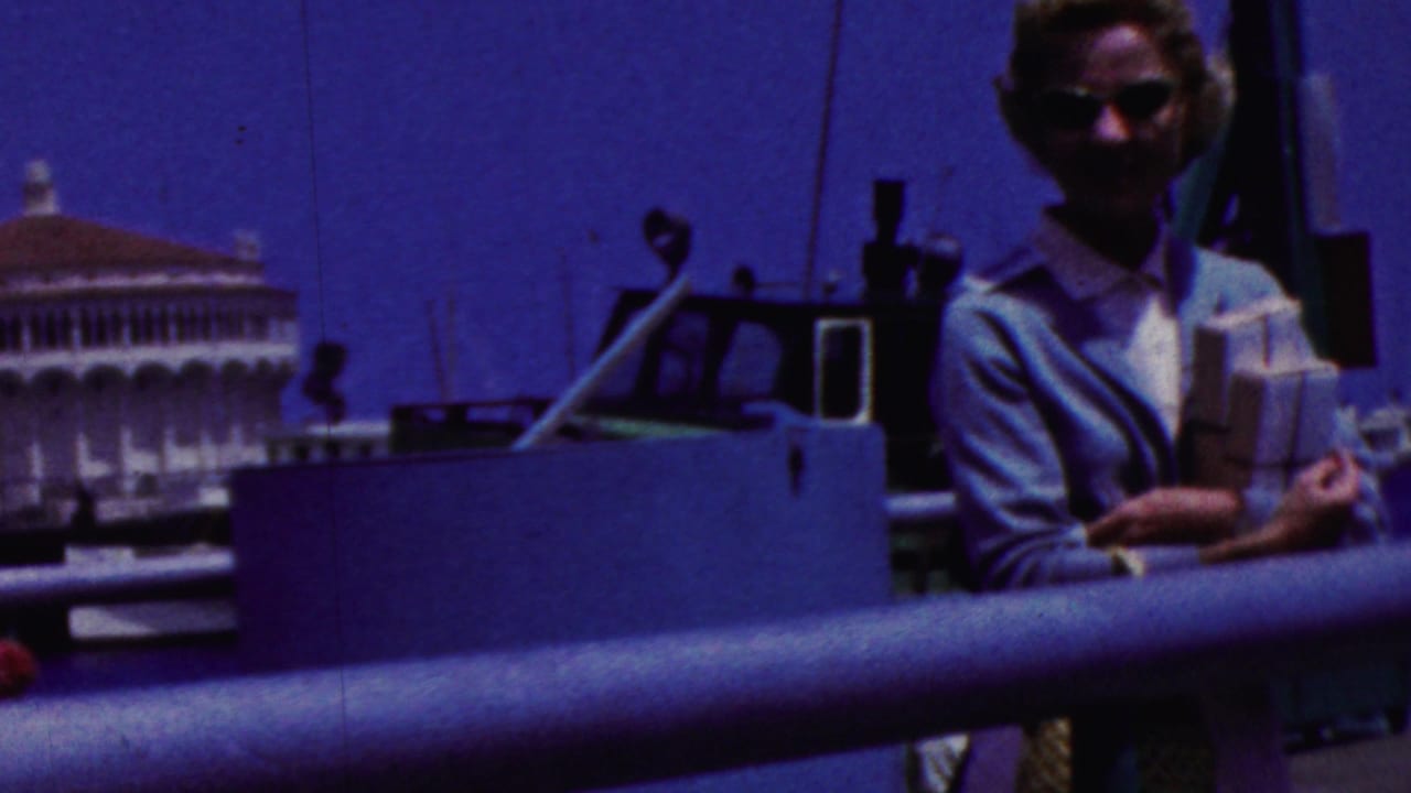 In 1961 California home-movie footage, a woman in sunglasses stands on a boat deck holding papers or books, with harbor water and a white waterfront building in the background.