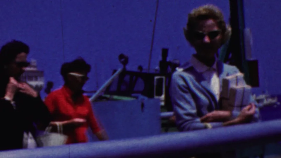 A home-movie frame shows the woman on the deck as other adult passengers enter the shot near the railing, with the harbor and white waterfront building still visible.