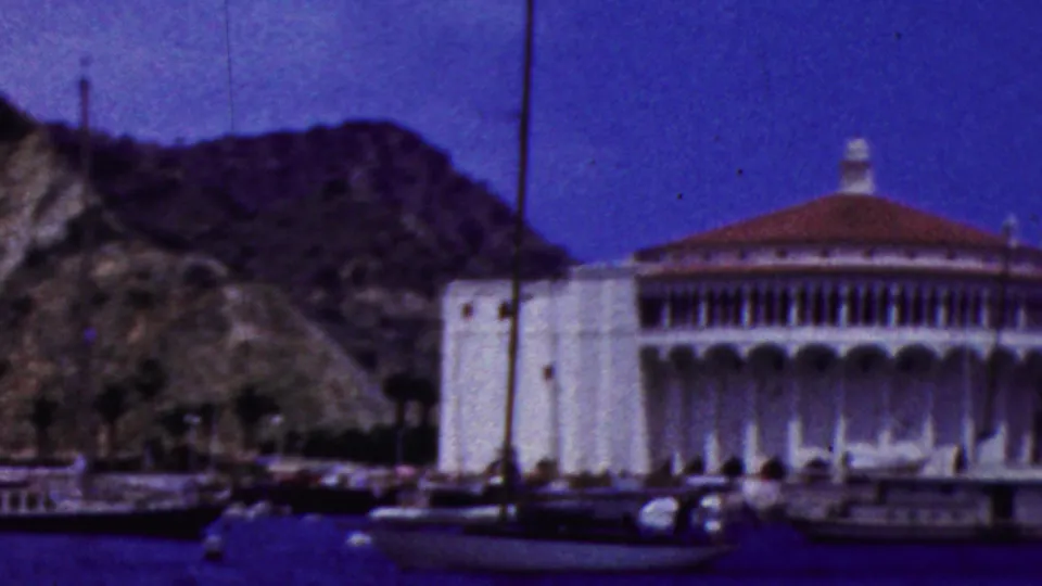 A white waterfront building stands beside a hillside and moored boats in the 1961 California coastal resort footage.