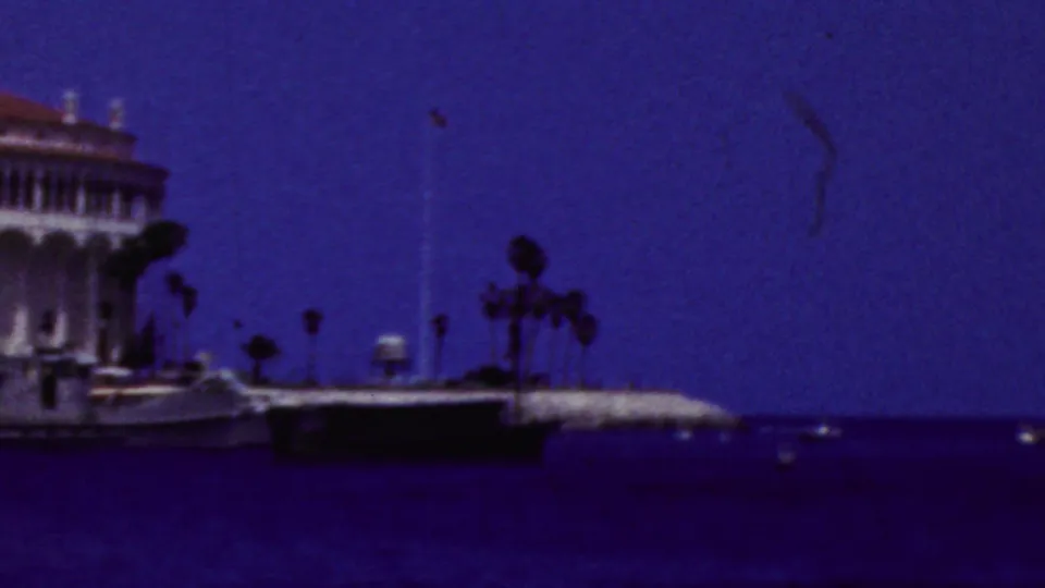 The camera has shifted past the main building, leaving part of the Catalina Casino at the frame edge with palm trees, pier structures, and deep blue water visible.