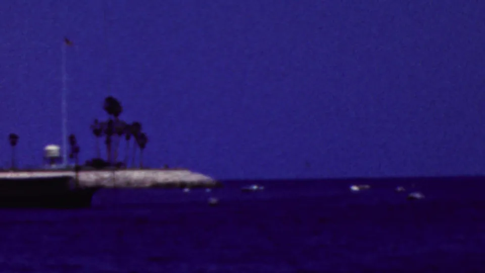 A later frame looks away from the casino toward open blue water, with a low pier or breakwater, palm trees, and distant small boats under a clear sky.