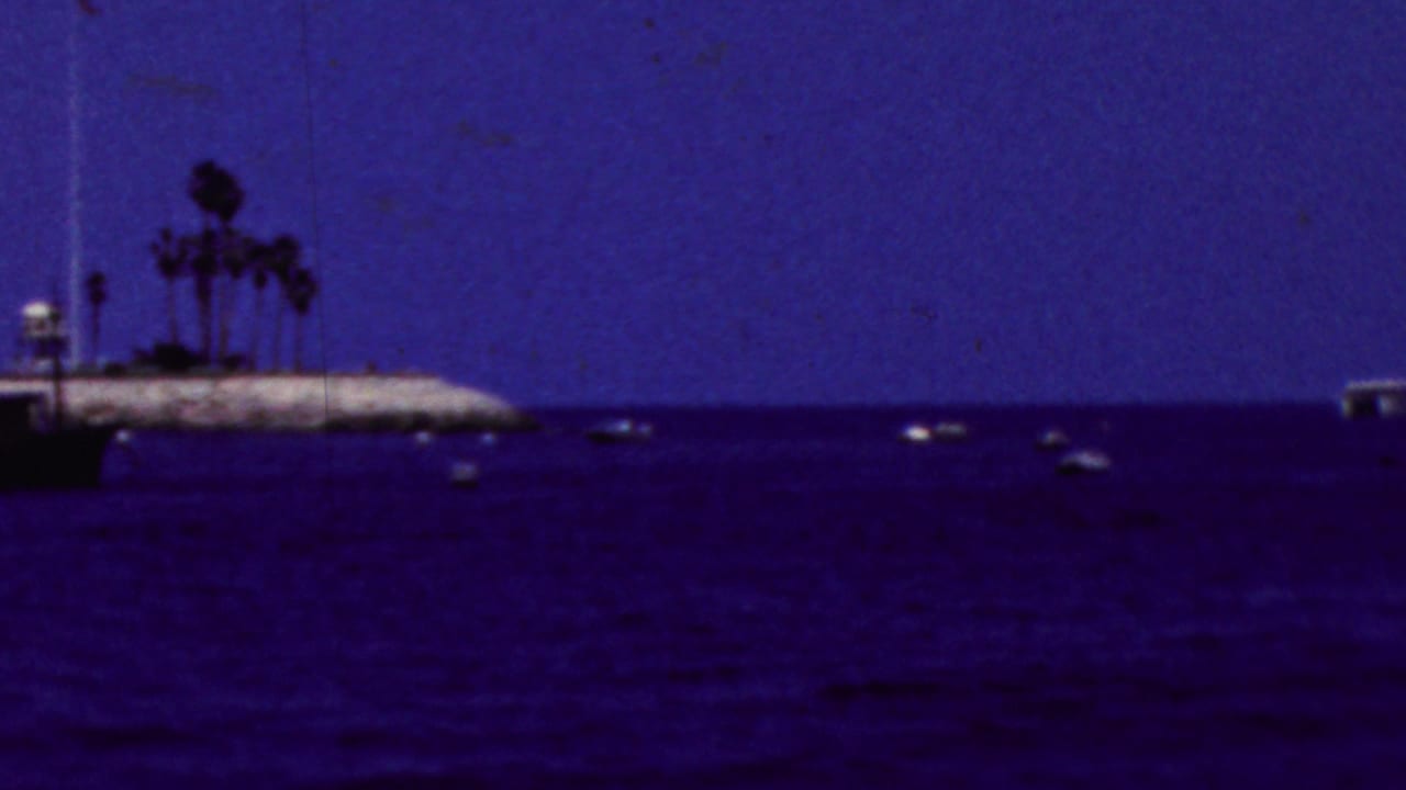 1961 California 8mm frame showing a dark blue bay, scattered small boats, and a rocky shoreline with palm trees in the distance.