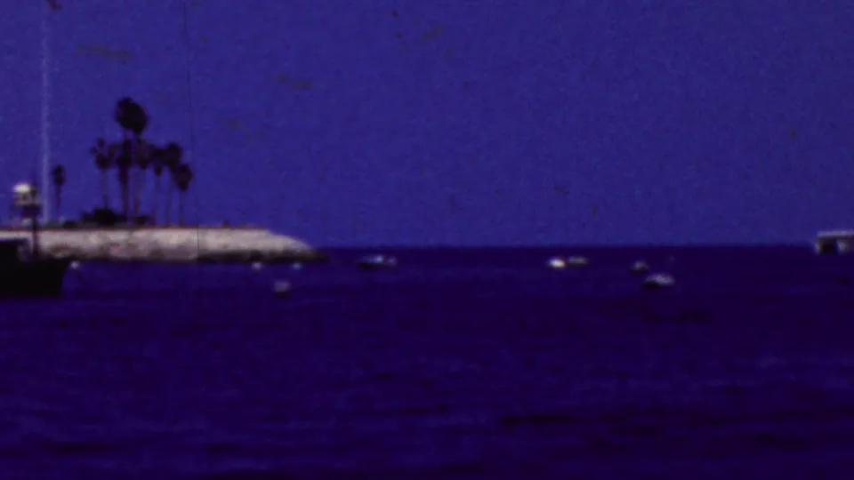 1961 California 8mm frame showing a dark blue bay, scattered small boats, and a rocky shoreline with palm trees in the distance.