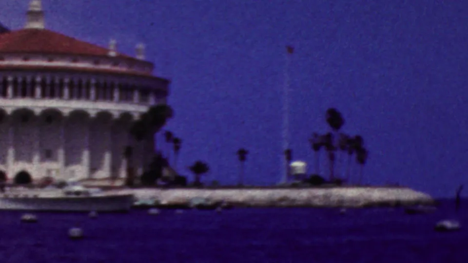 1961 California 8mm frame showing a white waterfront building with a red-tiled dome, small boats, palm trees, and blue water in front.