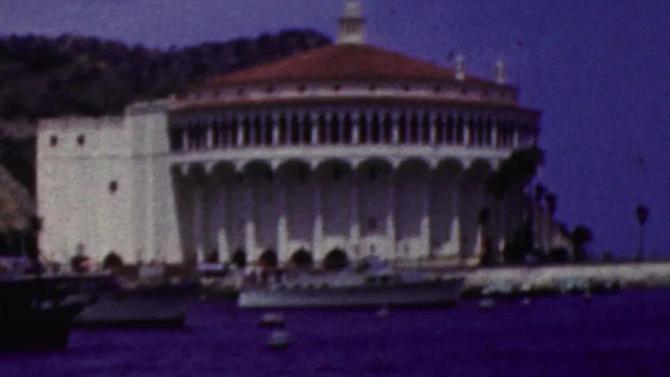 1961 California home-movie frame centered on a large white domed waterfront building with arched details, hillside behind it, and boats on the bay.