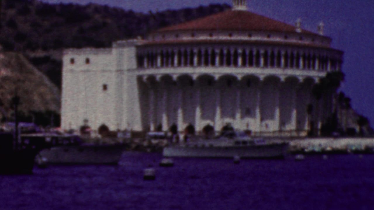 1961 California 8mm frame showing a white circular shoreline landmark with a red-tiled dome, arched facade, hillside backdrop, and dark blue harbor water in the foreground.