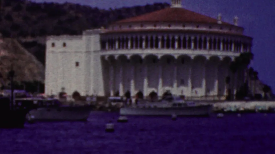 1961 California 8mm frame showing a white circular shoreline landmark with a red-tiled dome, arched facade, hillside backdrop, and dark blue harbor water in the foreground.