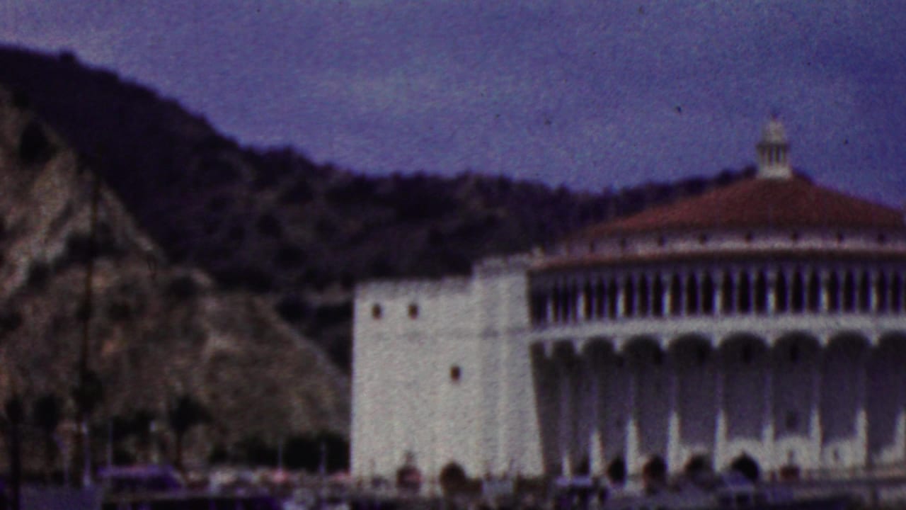 1961 California home-movie frame from a moving boat, with the white domed waterfront building shifted to the right against a rocky hillside and harbor water.