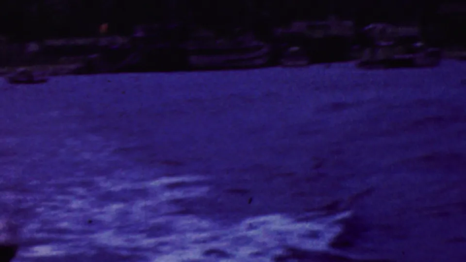 1961 California 8mm frame showing rippled harbor water, a dark shoreline band, small boats, and a partial view of the white waterfront landmark at upper right.