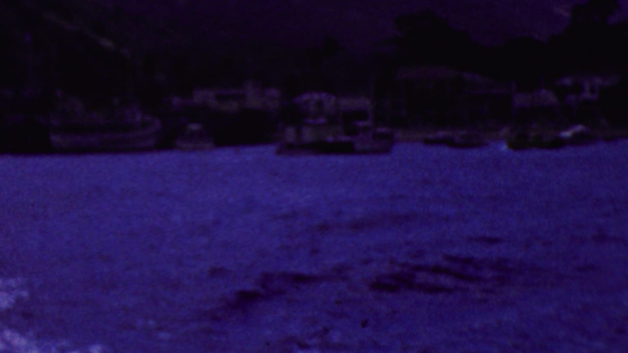 1961 California home-movie frame showing rippling water from a boat with a distant shoreline, houses, and moored small boats under heavy purple-blue color shift.
